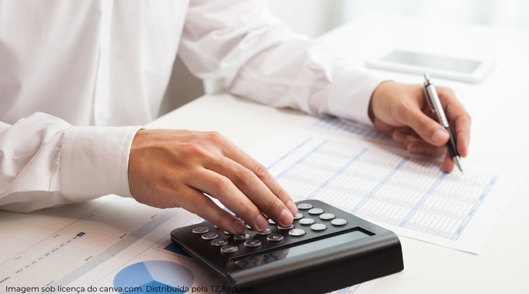 Mão esquerda segurando uma caneta e mão direita usando uma calculadora calculando quanto custa o seguro para agência de viagens.