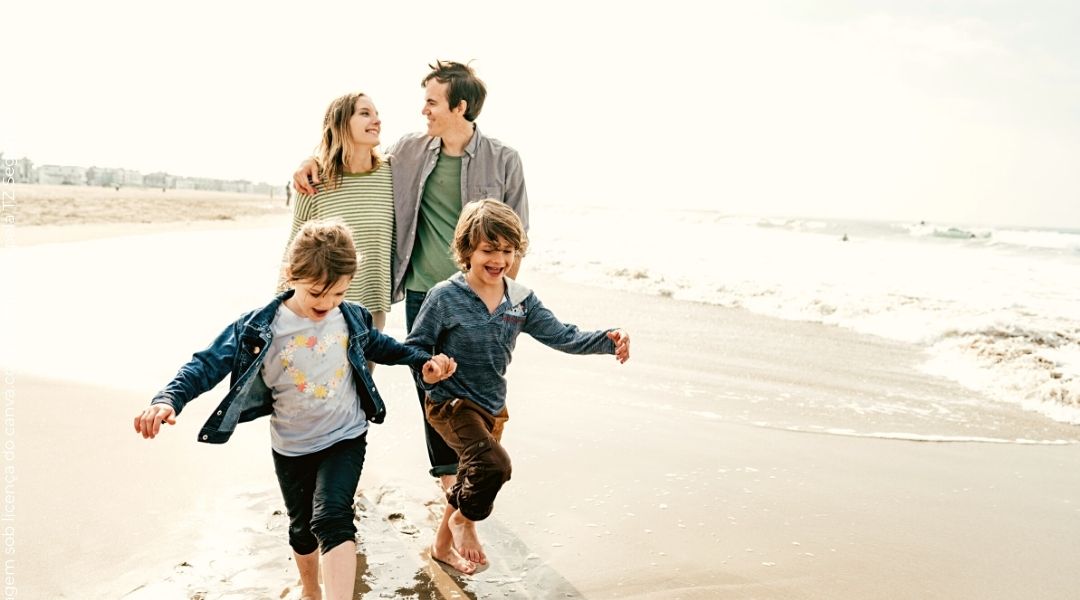 Família de 4 pessoas correndo em uma praia. Dois meninos correndo e brincando na frente, pai e mãe atrás sorrindo e abraçados