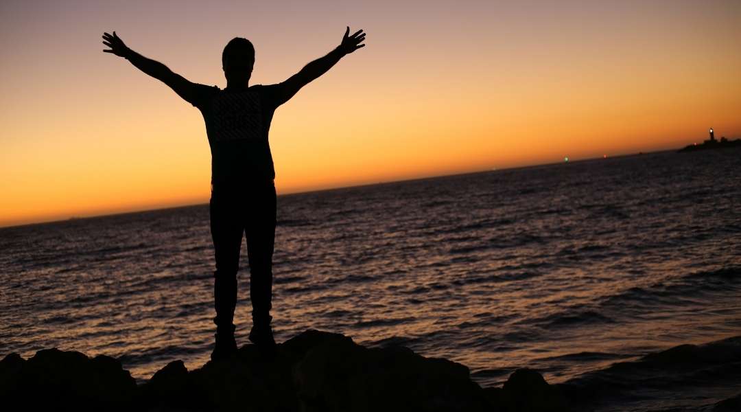 silhueta de homem jovem frente ao oceano no pôr do sol, ele esta de braços abertos, feliz com a vida