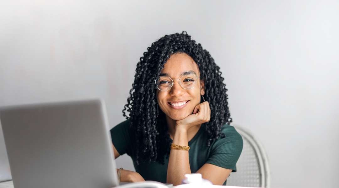 jovem mulher de óculos, cabelos cacheados e sorrindo, com o queixo apoiado na mão frente a um notebook.